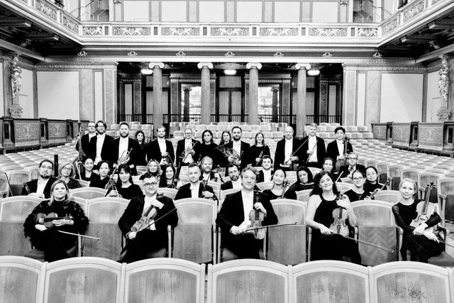 Black-and-white group portrait of Metropolitan Kammerorchester Wien musicians with their instruments in an ornate concert hall
